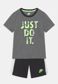 T-shirt grigio con la scritta "JUST DO IT" in lettere verdi e argento, abbinato a pantaloni corti neri con il logo Nike verde e accenti bianchi.