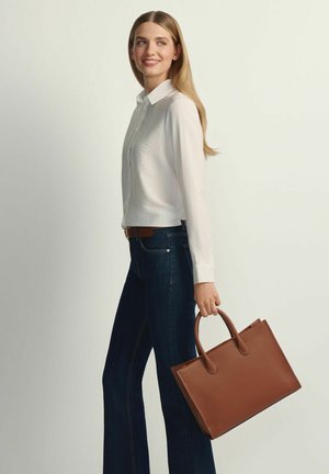 Kobieta w białej bluzce i ciemnych dżinsach, trzymająca dużą brązową skórzaną torebkę, patrząca na bok z delikatnym uśmiechem.