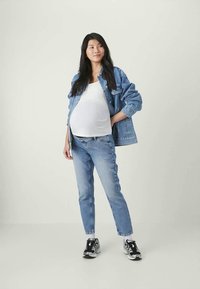 Mulher grávida vestindo uma blusa branca, jaqueta de ganga, jeans e ténis, segurando uma das mãos na anca contra um fundo simples.
