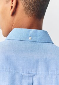 Lichtblauw chambray overhemd met een button-down kraag, een plooi aan de achterkant en een klein knoopdetail aan de achterkant van de kraag. Gladde textuur.