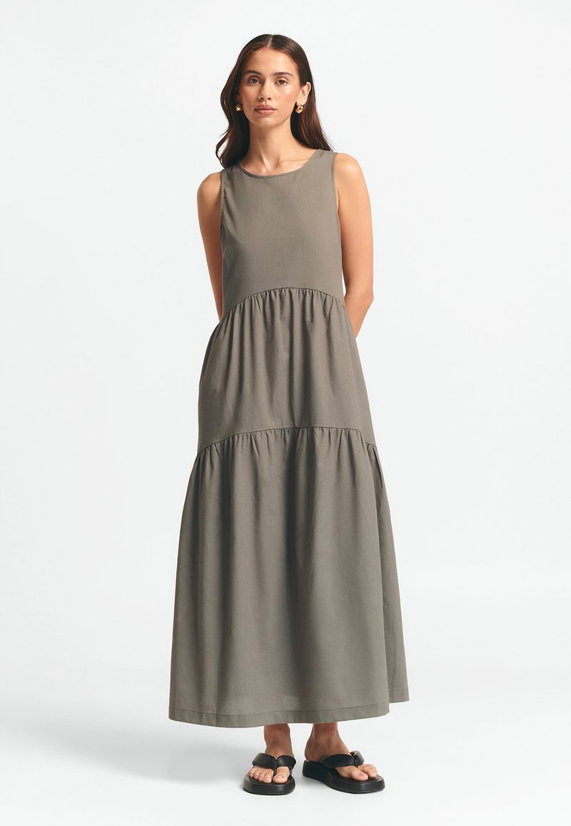 Ilga, be rankovių maxi suknelė alyvuogių spalvos, su apvaliu kaklu ir daugiapakopių volanų apdaila. Dėvima su juodais šlepetėmis.