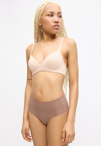 Jeune femme aux longs tresses blondes vêtue d'un soutien-gorge nude et d'une culotte taille haute mauve, regardant sur le côté contre un fond blanc.