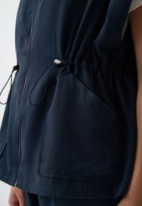 Gilet léger marine avec taille élastique à cordon de serrage, deux poches latérales et texture lisse. Doté d'une fermeture éclair frontale intégrale et d'une coupe décontractée.