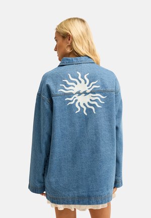 Femme aux cheveux blonds portant une veste en jean bleue avec un motif de soleil blanc dans le dos et une robe blanche en dessous, tournée dos à la caméra.