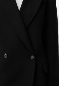 Svart dubbelknäppt kavaj gjord av ullblandning med slagna lapeller, som har två svarta knappar och en bröstficka på vänster sida.