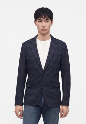 Jeune homme portant un blazer à carreaux bleu foncé sur un t-shirt blanc et un jean bleu, debout devant un fond blanc uni.