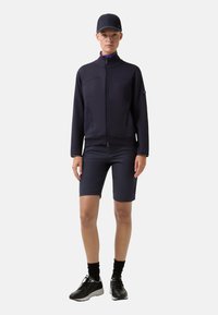 Námořnicky modrý sportovní komplet zahrnující bundu na zip s vysokým límcem a odpovídající šortky. Stylizováno s černými ponožkami a teniskami.