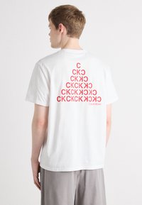 White cotton t-shirt featuring a red graphic pattern on the back, displaying repeated letters “CK” in varying sizes, with Calvin Klein logo.