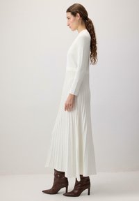 Vrouw met lang gevlochten haar draagt een witte geribbelde jurk met lange mouwen en bruine puntige hoge hakken, staande in profiel tegen een effen achtergrond.
