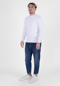 Felpa bianca a maniche lunghe con dettagli a costina, abbinata a jeans in denim blu. Il modello indossa scarpe da ginnastica bianche, in piedi di fronte a uno sfondo semplice.