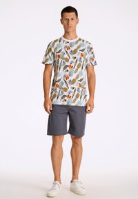 T-shirt med färgglada frukt- och växtmönster på en vit bakgrund, ihop med grå shorts och vita sneakers. Avslappnad och lätt design.