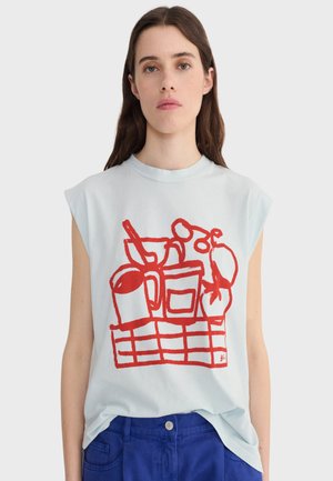 Mladá žena v bielom tielku bez rukávov s červeným abstraktným čiarovým obrázkom kuchynských predmetov na prednej strane a modrých nohaviciach.