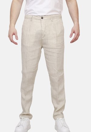 Uomo che indossa pantaloni di lino beige, sneakers bianche e una camicia bianca infilata, in piedi davanti a uno sfondo semplice.