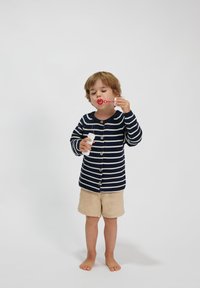 Gestreifter navyblauer und cremfarbener Cardigan mit Knöpfen, beiger Shorts und Seifenblasen in der Hand. Kind steht barfuß vor einem weißen Hintergrund.