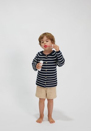 Gestreifter navyblauer und cremfarbener Cardigan mit Knöpfen, beiger Shorts und Seifenblasen in der Hand. Kind steht barfuß vor einem weißen Hintergrund.