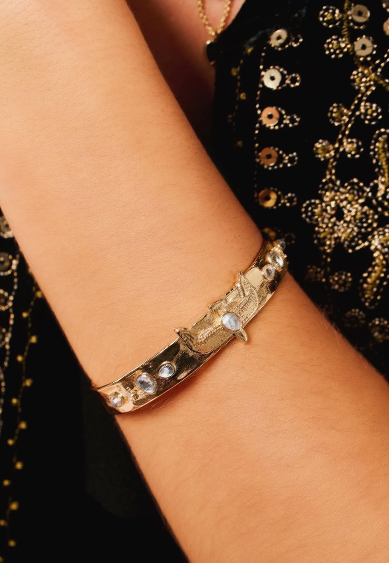 Gold-tone bracelet featuring ornate design and blue gemstones. Smooth, sleek band with textured accents and central decorative element.