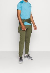 En man som bär en blå Under Armour t-shirt, olivgröna joggers, grå sneakers och en olivgrön sweatshirt knuten runt midjan.