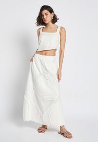 Weißes, kurzes Tanktop mit quadratischem Ausschnitt, kombiniert mit einem langen, fließenden Rock, der strukturierte, gestickte Muster und ausgefranste Ränder aufweist.