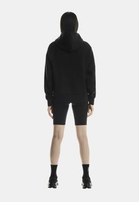 Schwarzer Kapuzenpulli mit Kapuze, Raglanärmeln und gerippten Bündchen, kombiniert mit schwarzen Radshorts und schwarzen Sportschuhen, von hinten fotografiert.