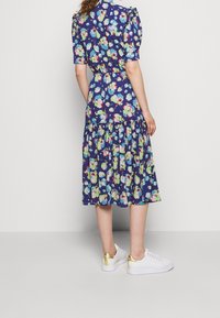 Květinové šaty s krátkými nabíranými rukávy, přiléhavým předním dílem a vrstevnatou sukní v námořnické modré barvě s barevnými květinovými vzory. Nosené s bílými teniskami.