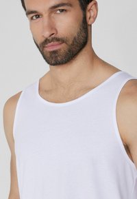 Top de algodón blanco sin mangas con escote redondeado y tirantes anchos, textura suave y diseño ajustado. Primer plano de la parte superior del cuerpo del modelo.