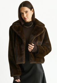 Woman wearing a dark brown faux fur jacket over a brown turtleneck and black satin skirt, with gold hoop earrings and a chain necklace.