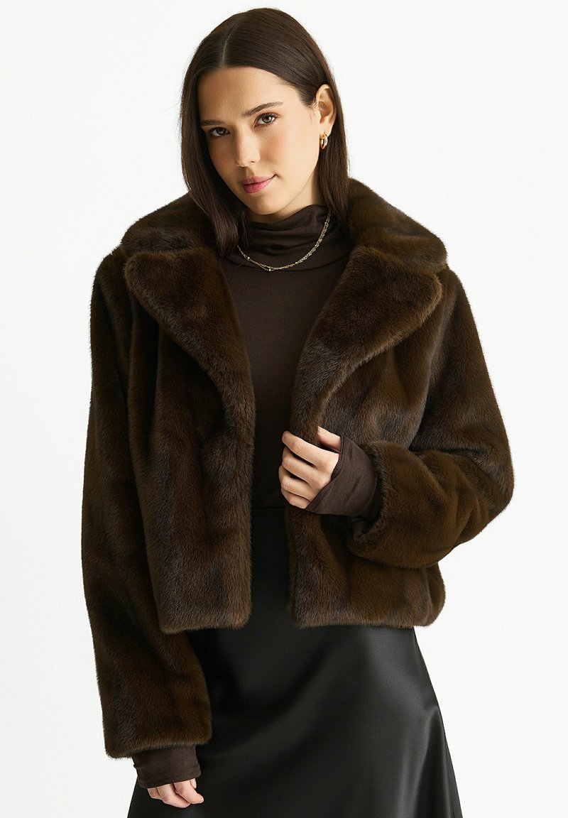 Woman wearing a dark brown faux fur jacket over a brown turtleneck and black satin skirt, with gold hoop earrings and a chain necklace.