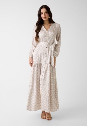 Femme aux longs cheveux bruns portant une robe maxi beige à manches longues, boutonnée, avec une ceinture assortie et des talons ouverts, posant devant un fond blanc.