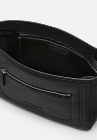 Czarna skórzana torebka o gładkiej fakturze, z zamkiem błyskawicznym na górze i kieszonką zewnętrzną, z podkreślonymi przeszyciami oraz markową metką wewnętrzną.