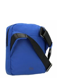 Bolso de hombro de tela azul con forma rectangular, correa ajustable negra y cierre de cremallera. Exterior texturizado y detalle sutil del logo.