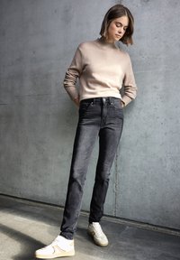 Suéter de punto beige claro con un cuello alto, combinado con jeans semi ajustados negros y zapatillas blancas con detalles beige, frente a un fondo de concreto.