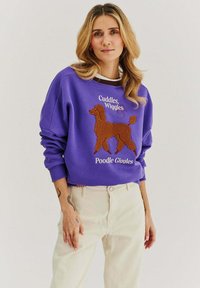 Fialová mikina s texturovaným hnědým designem pudla a bílým textem, s žebrovanými manžetami a volným střihem, spárovaná se světlými kalhotami.