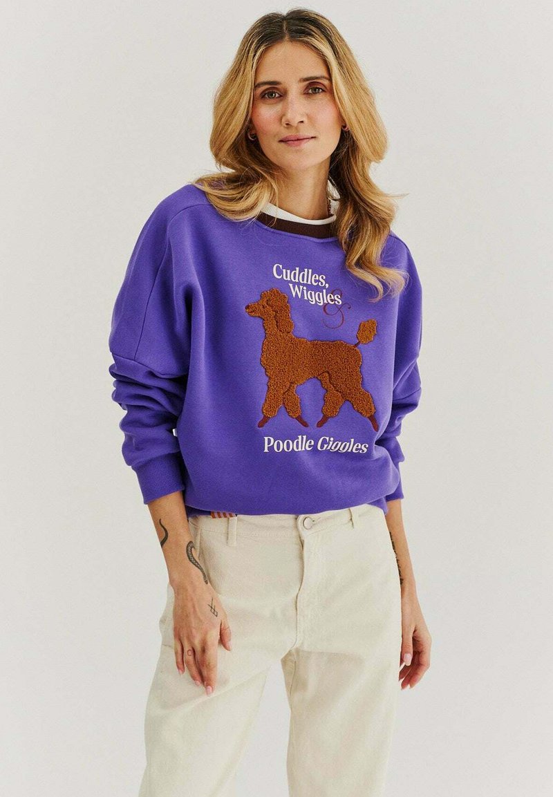 Fialová mikina s texturovaným hnědým designem pudla a bílým textem, s žebrovanými manžetami a volným střihem, spárovaná se světlými kalhotami.