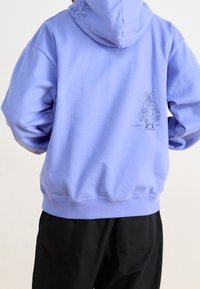 Ljust lila hoodie med en känguruficka och ribbade muddar. Har en linjetecknad figur på ryggen och ett mönstrat detaljer på huvan.