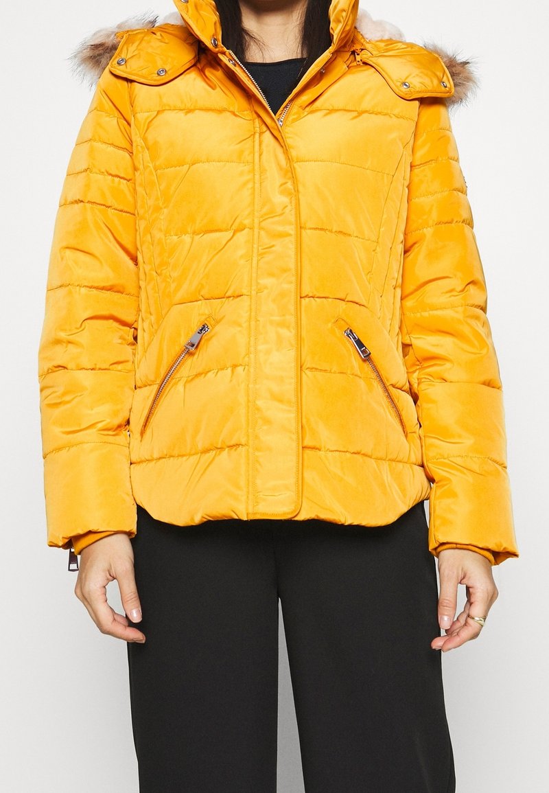 Giubbotto giallo imbottito con cappuccio e trim in pelliccia sintetica. Design trapuntato, due tasche laterali con zip e chiusura frontale con zip.