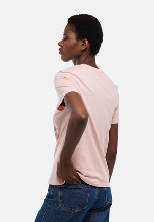 Femme aux cheveux courts et bouclés, portant un t-shirt rose clair et un jean bleu, debout de profil avec une main sur la hanche.