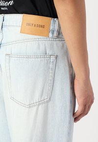 Light blue denim jeans with a relaxed fit, featuring a tan leather label with "ONLY & SONS" on the waistband, and a back pocket detail.