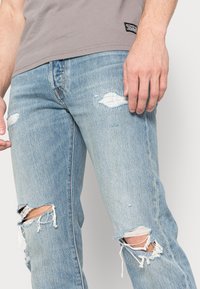 Ljusblå denimjeans med slitna hål vid knäna, rak passform och fransade kanter. Vita sömmar och en subtil etikett på midjebandet.