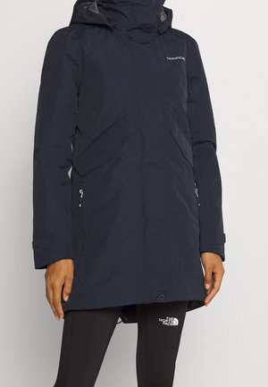 Person trägt eine lange navyblaue Didriksons Kapuzenjacke mit Reißverschluss und Klappentaschen, kombiniert mit schwarzen Leggings von The North Face.