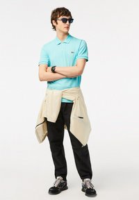 Polo azzurro chiaro, pantaloni neri, giacca beige legata in vita, occhiali da sole e scarpe da ginnastica; con logo Lacoste e tessuti strutturati.