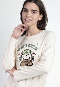 Camisa de manga larga color crema con un gráfico de un personaje verde con orejas grandes y un libro, acompañado del texto "LA FUERZA ES FUERTE."