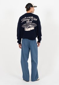 Homme portant une casquette noire, un pull bleu marine avec l'inscription "In a rush Racing Club Berlin" et un motif de voiture, ainsi qu'un pantalon bleu ample, se tenant devant un fond blanc.