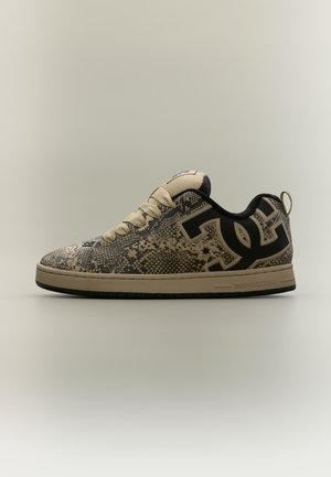 Χαμηλό sneaker με μπεζ σχέδιο φίδι, μαύρο λογότυπο DC στο πλάι, μπεζ κορδόνια και μπεζ σόλα με μαύρο κάτω μέρος.
