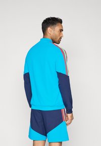 Világoskék melegítőfelső sötétkék ujjakkal, a hátoldalán piros, sárga és zöld három függőleges csíkkal.