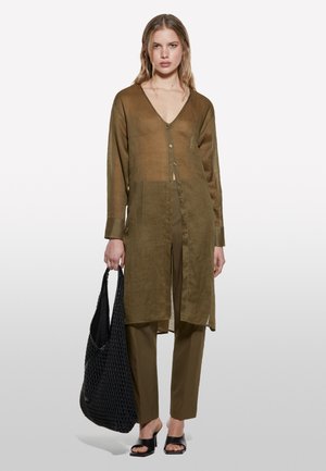 Chemise transparente vert olive à boutons et manches longues, accompagnée d'un pantalon assorti. Le modèle tient un tote noir en osier et porte des sandales noires à enfiler.