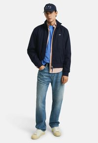 Giubbotto bomber blu sopra una maglietta a righe rosa chiaro, con una t-shirt blu sotto, jeans azzurri e sneaker color crema. Cappellino nero con logo.