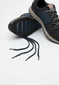 Scarpe da trail di colore blu scuro e grigio con suola testurizzata, parte superiore in mesh e accenti riflettenti. I lacci sono allentati e mostrati in un'applicazione piatta.