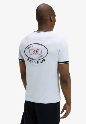 Homme de dos portant une chemise à manches courtes bleu clair avec bordure noire et grand logo Eden Park avec nœud rose au dos.