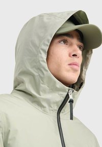 Hellgrüne wasserdichte Jacke mit hohem Kragen, Reißverschluss und verstellbarer Kapuze. Der Stoff wirkt glatt und leicht reflektierend.