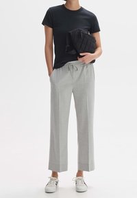 T-shirt noir à manches courtes associé à un pantalon large gris clair et des baskets blanches. Un sac bandoulière noir matelassé repose sur la hanche.